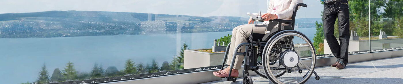 Layout Bild Seitenkopf Rollstuhl mit E-Fix Zusatzantrieb und mit einer Person